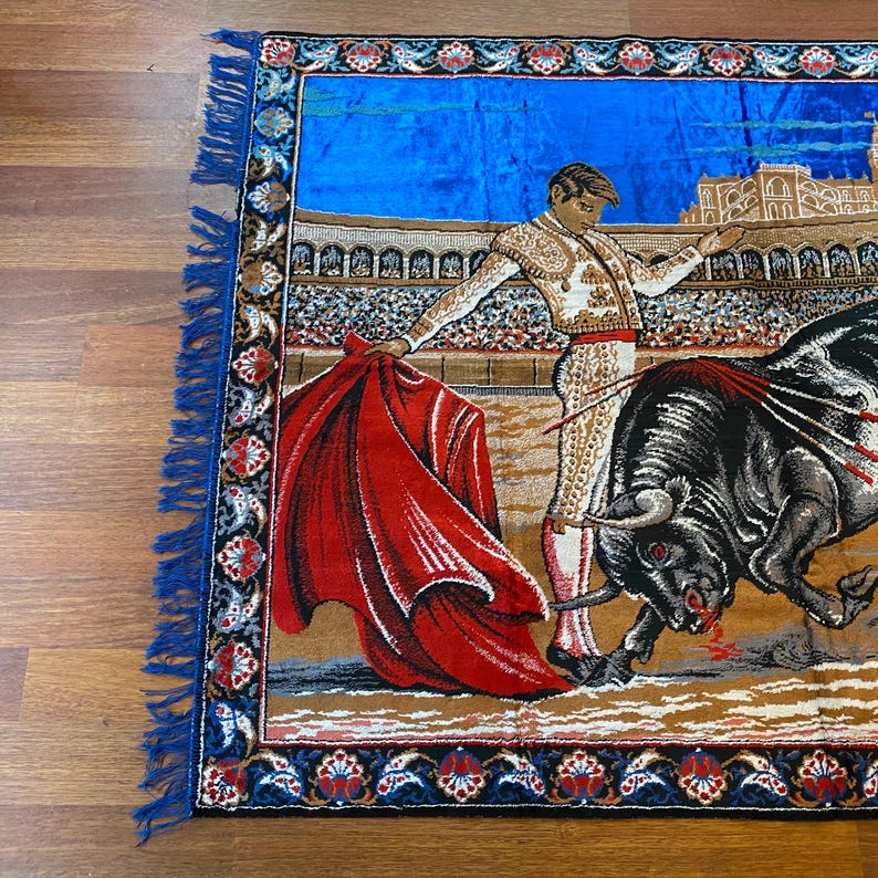 K&ouml;nnte beinhalten: Wandteppich mit einer Stierszenen-Darstellung. Ein Matador in traditioneller Kleidung steht einem angreifenden schwarzen Stier mit rotem Umhang gegen&uuml;ber. Der Hintergrund zeigt ein Stadion und blauen Himmel, mit dekorativem Rand und blauer Fransen.