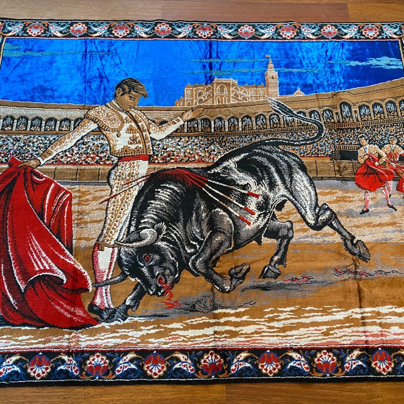 K&ouml;nnte beinhalten: Wandteppich mit einer Stierszenen-Darstellung. Ein Matador in einem gold-wei&szlig;en Anzug steht einem schwarzen Bullen mit roten Akzenten gegen&uuml;ber. Der Hintergrund zeigt ein Stadion mit Zuschauern und blauem Himmel.