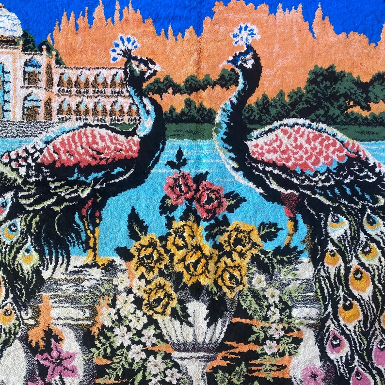 Peut inclure: Textile d&eacute;coratif avec deux paons aux plumes &eacute;labor&eacute;es, sur fond de palais, d'eau et de fleurs. Les paons ont un plumage noir, rouge et bleu. Un vase de fleurs jaunes et roses est au premier plan.