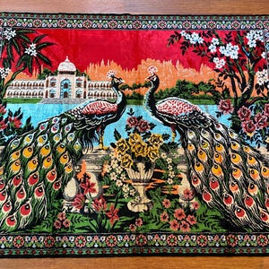 May include: A decorative rug depicting two peacocks with detailed, colourful feathers. The rug features a scenic background with a building, water, and floral designs. The border has a floral pattern and red fringe.