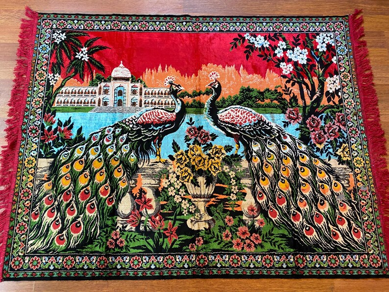 May include: A decorative rug depicting two peacocks with detailed, colourful feathers. The rug features a scenic background with a building, water, and floral designs. The border has a floral pattern and red fringe.