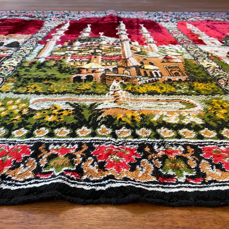 May include: A detailed prayer rug with a Middle Eastern city scene, including minarets and buildings. The rug has a red border and a black fringe. The design incorporates green, yellow, and brown colours. The rug is likely made of woven fabric.