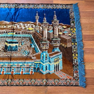 May include: A prayer rug featuring a detailed illustration of a religious site with minarets and buildings. The rug has a blue border with fringe and a brown and blue central design. The rug is rectangular.