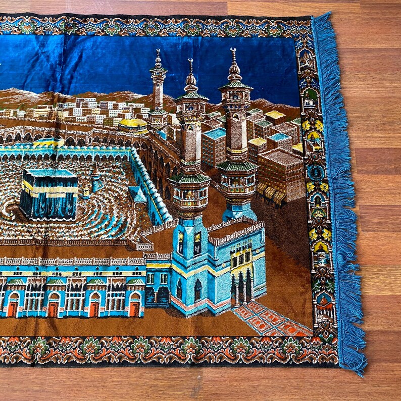 May include: A prayer rug featuring a detailed illustration of a religious site with minarets and buildings. The rug has a blue border with fringe and a brown and blue central design. The rug is rectangular.