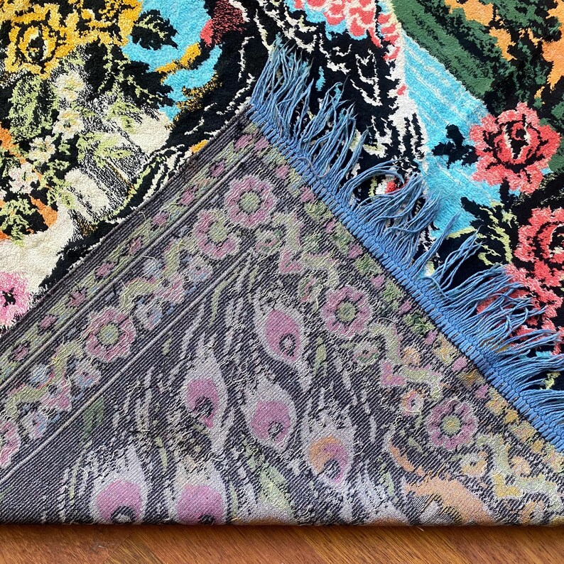 Peut inclure: Un tapis &agrave; motifs floraux color&eacute;s sur fond noir. Le tapis pr&eacute;sente diverses fleurs dans des tons de rose, jaune et bleu. Il poss&egrave;de une frange bleue et une bordure d&eacute;taill&eacute;e avec des motifs floraux et de plumes de paon.