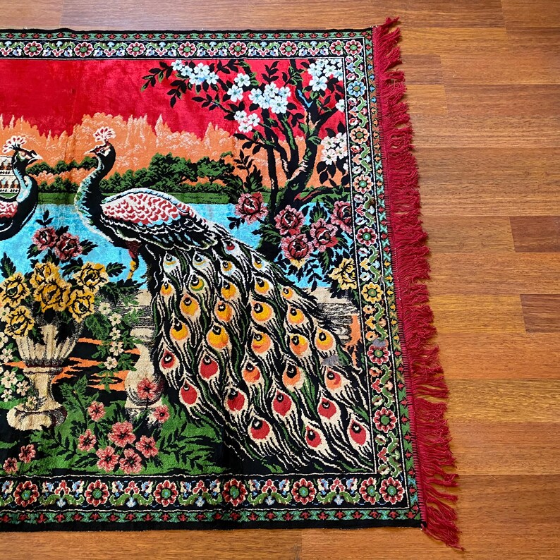 May include: A rectangular rug with a peacock design. The rug has a red background with a landscape scene, including peacocks, flowers, and trees. The border is black with floral patterns, and the edge has red fringe.