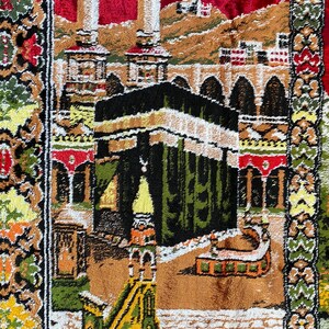 May include: A detailed prayer rug with a central depiction of the Kaaba in Mecca. The rug features a complex design with architectural elements, including minarets and arches, in shades of green, brown, red, and gold.