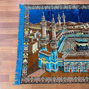 May include: A prayer rug with a detailed illustration of a religious site, featuring minarets and buildings in shades of blue, brown, and gold. The rug has a blue border with fringe.