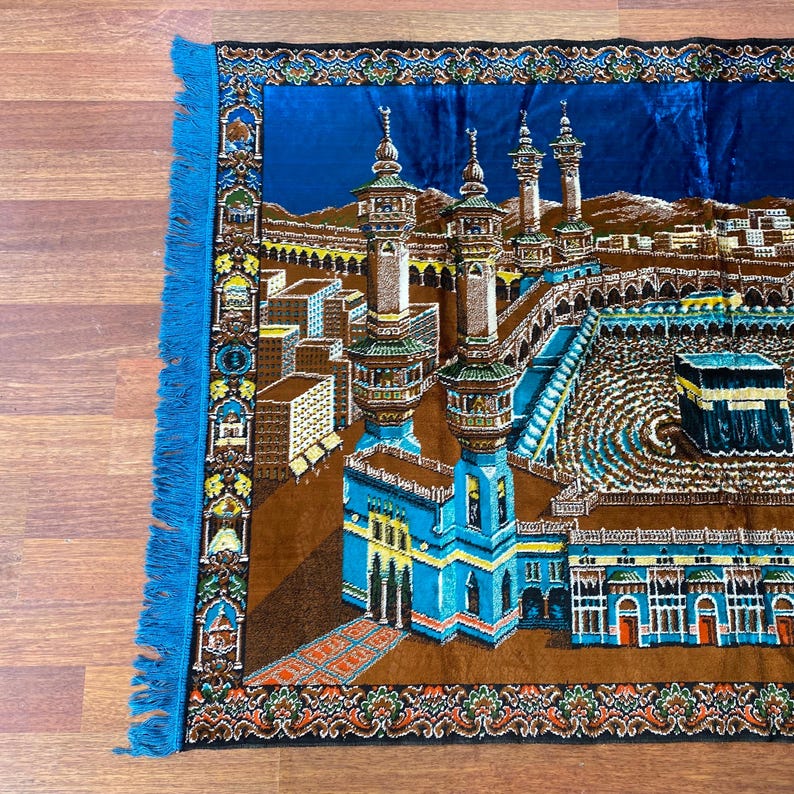 May include: A prayer rug with a detailed illustration of a religious site, featuring minarets and buildings in shades of blue, brown, and gold. The rug has a blue border with fringe.