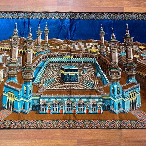 May include: A rectangular prayer rug featuring a detailed illustration of the Masjid al-Haram in Mecca. The rug has a blue background with a central image of the Kaaba, surrounded by minarets and buildings. The rug has a brown border with blue fringe.