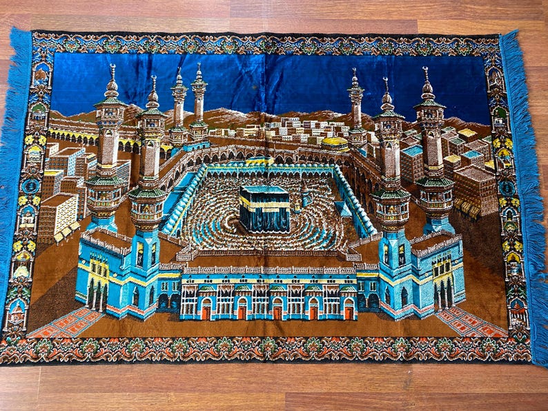 May include: A rectangular prayer rug featuring a detailed illustration of the Masjid al-Haram in Mecca. The rug has a blue background with a central image of the Kaaba, surrounded by minarets and buildings. The rug has a brown border with blue fringe.