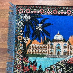 Peut inclure: Tapis d&eacute;coratif rectangulaire bleu avec une illustration d&eacute;taill&eacute;e d'un palais, d'un palmier et de plumes de paon. Le tapis a une bordure florale et des franges sur les bords. La palette de couleurs comprend le bleu, l'orange, le vert et le blanc.