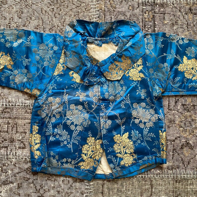May include: A blue silk jacket with a gold floral pattern and a large collar. The jacket has a button closure and is in good condition.
