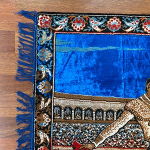 K&ouml;nnte beinhalten: Rechteckiger Teppich mit blauem Samtmittelteil und dekorativem Rand. Der Rand weist florale und geometrische Muster in Rot, Schwarz und Gold auf. Der Teppich hat eine Franse auf einer Seite und zeigt einen Stierk&auml;mpfer.