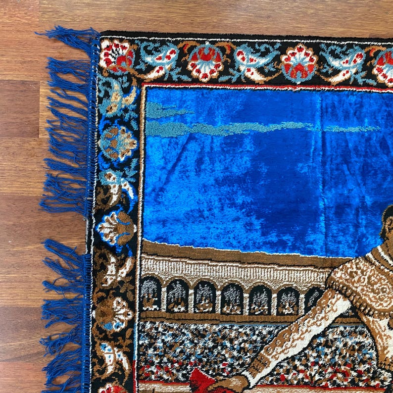 K&ouml;nnte beinhalten: Rechteckiger Teppich mit blauem Samtmittelteil und dekorativem Rand. Der Rand weist florale und geometrische Muster in Rot, Schwarz und Gold auf. Der Teppich hat eine Franse auf einer Seite und zeigt einen Stierk&auml;mpfer.