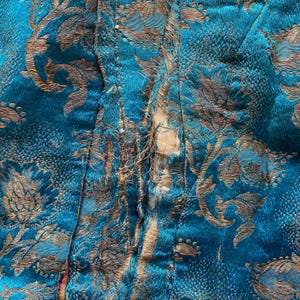 Puede incluir: Primer plano de una tela azul turquesa con un patr&oacute;n floral dorado. La tela est&aacute; rasgada a lo largo de la costura, revelando los bordes sin terminar.