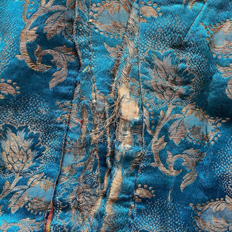 Puede incluir: Primer plano de una tela azul turquesa con un patr&oacute;n floral dorado. La tela est&aacute; rasgada a lo largo de la costura, revelando los bordes sin terminar.