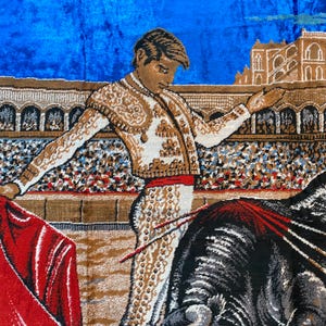 K&ouml;nnte beinhalten: Ein Wandteppich, der eine Stierszenen zeigt. Ein Matador in einem dekorativen Anzug steht in einer Arena mit einem roten Umhang und blickt einem schwarzen Stier entgegen. Der Hintergrund zeigt einen blauen Himmel und ein Stadion.