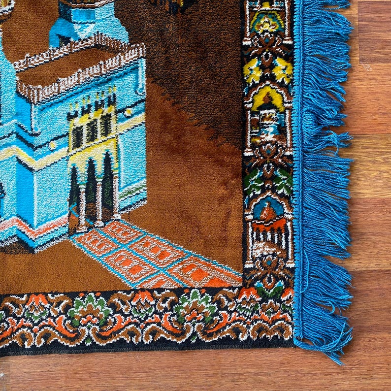 May include: A prayer rug with a detailed design featuring a mosque in shades of blue and brown. The rug has a brown background with an orange and blue geometric pattern and a blue fringe.