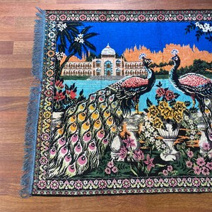 Peut inclure: Tapis color&eacute; avec une illustration d&eacute;taill&eacute;e de deux paons, d'un palais et de jardins luxuriants. Le tapis a un fond bleu avec une bordure florale et des bords frang&eacute;s. Les paons ont un plumage vibrant avec des accents roses, jaunes et verts.