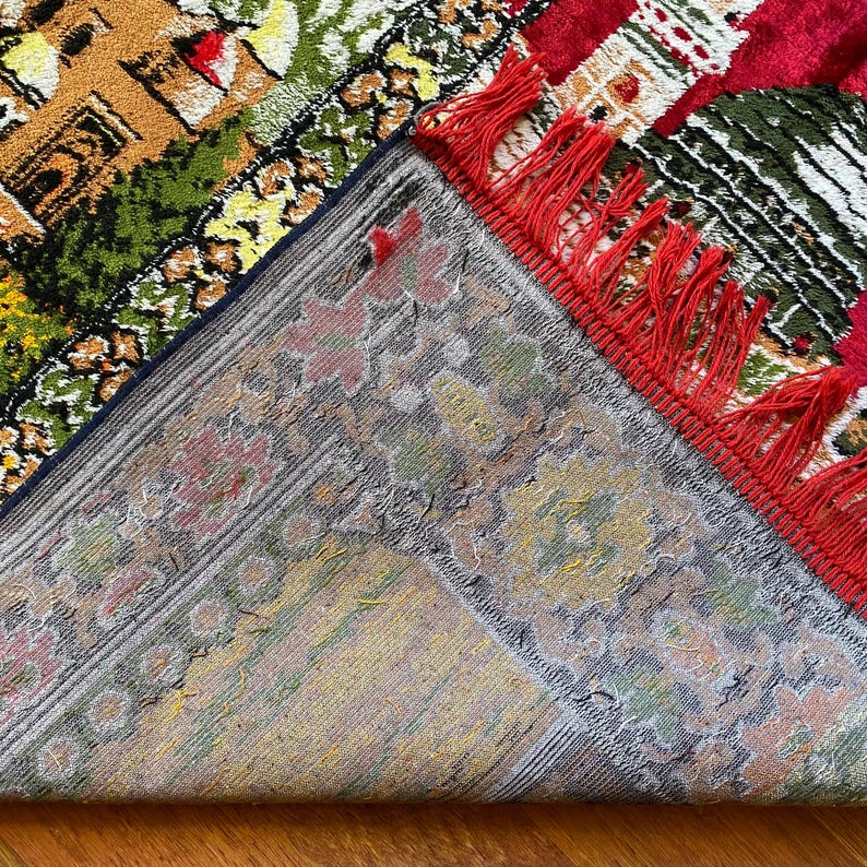 May include: A colourful, vintage rug with a detailed design. The rug features a red border with red fringe, and a pattern of buildings, trees, and floral elements in shades of green, yellow, and brown. The underside of the rug is visible.