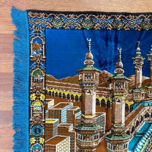 May include: A blue prayer rug with a detailed illustration of a religious site. The rug features a central image of a building with minarets, surrounded by a border with floral patterns and blue fringe.