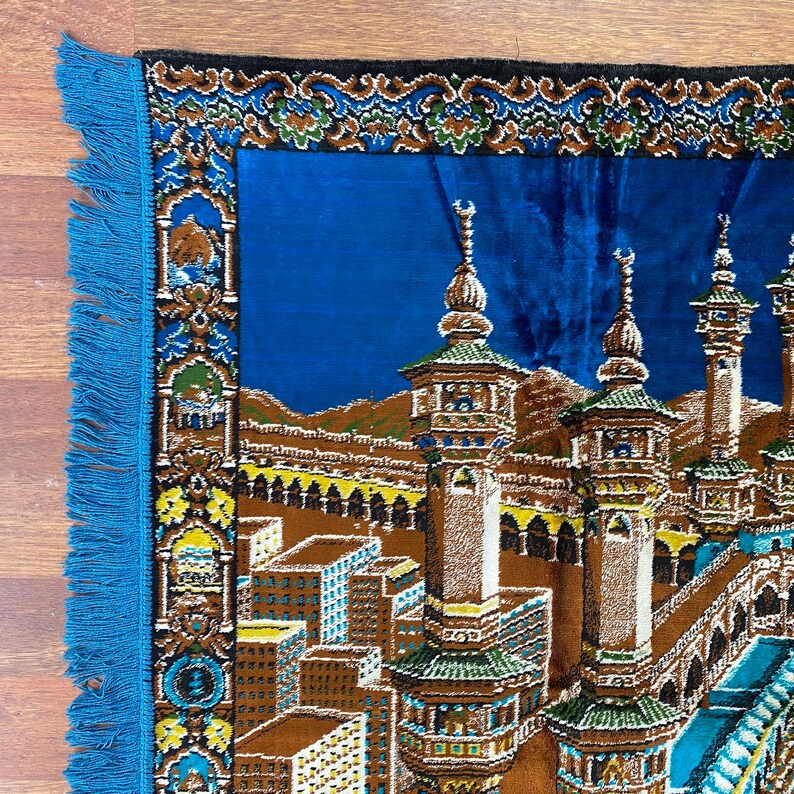 May include: A blue prayer rug with a detailed illustration of a religious site. The rug features a central image of a building with minarets, surrounded by a border with floral patterns and blue fringe.