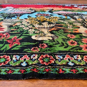 May include: A close-up of a vibrant rug with a floral and bird design. The rug has a black background with green, red, and blue floral patterns. A large vase of yellow flowers is in the center, with birds on either side.