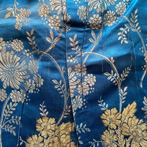 May include: Close-up of a blue fabric with a gold floral pattern. The fabric has a subtle sheen and a textured surface.