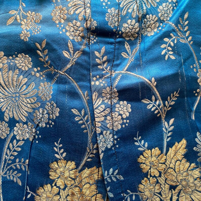 May include: Close-up of a blue fabric with a gold floral pattern. The fabric has a subtle sheen and a textured surface.