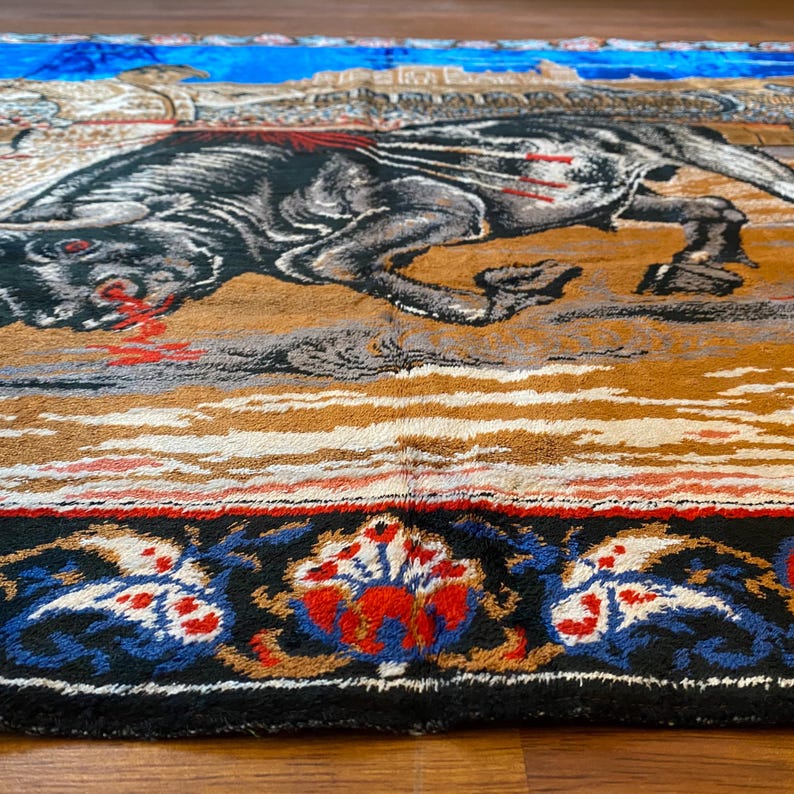 K&ouml;nnte beinhalten: Ein detaillierter Wandteppich mit einer schwarzen und grauen Tierfigur, m&ouml;glicherweise einem Pferd oder Bullen, mit roten Akzenten. Der Hintergrund umfasst einen blauen Himmel und eine braune Landschaft. Der Rand hat eine schwarze Basis mit blauen, roten und wei&szlig;en Blumenmustern.