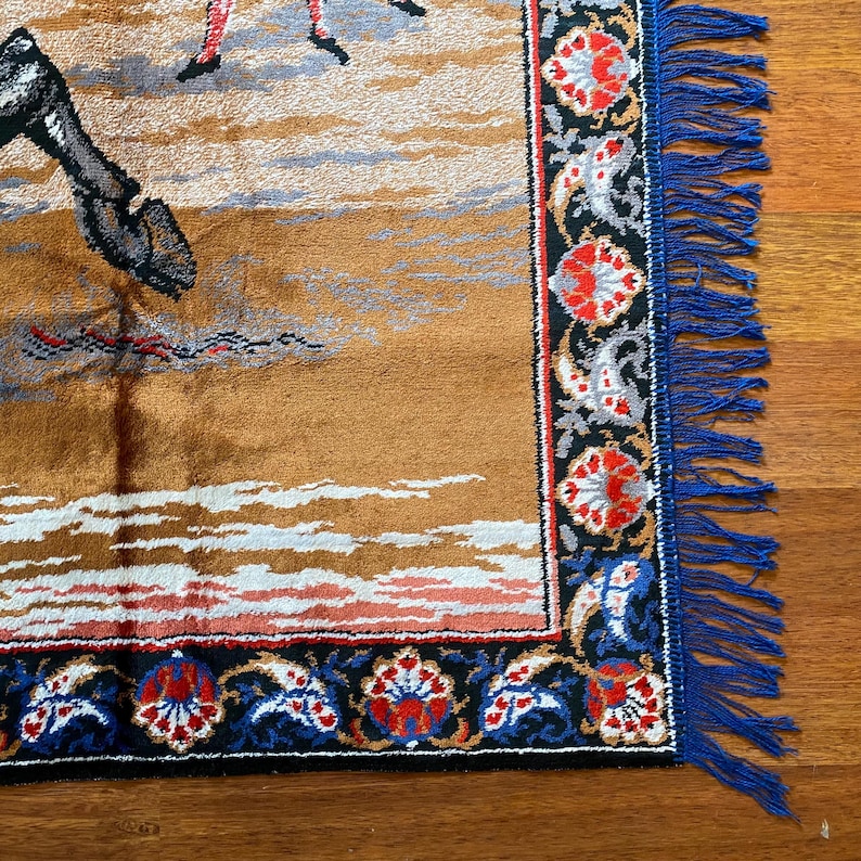 K&ouml;nnte beinhalten: Ein rechteckiger Wandteppich mit einer braunen und beigen Landschaftsszene, mit Figuren in Rot und Schwarz. Der Rand ist schwarz, blau und rot mit floralen Mustern, und der Rand hat blaue Fransen.