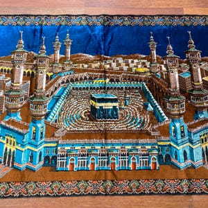 May include: A detailed prayer rug featuring a depiction of the Kaaba in Mecca. The rug showcases a central black cube, surrounded by a courtyard and minarets, with a blue sky background and ornate borders. The color palette includes shades of blue, brown, and gold.