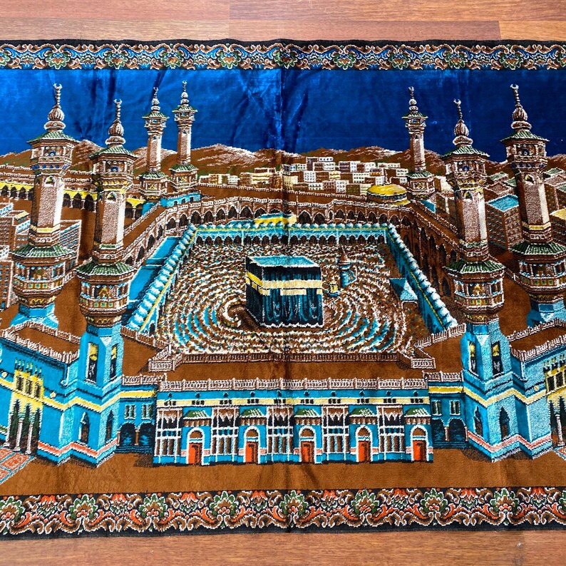May include: A detailed prayer rug featuring a depiction of the Kaaba in Mecca. The rug showcases a central black cube, surrounded by a courtyard and minarets, with a blue sky background and ornate borders. The color palette includes shades of blue, brown, and gold.