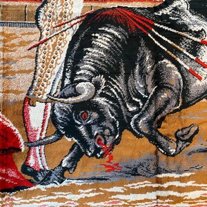 K&ouml;nnte beinhalten: Ein Wandteppich, der eine Stierszenen darstellt. Ein schwarzer Stier mit grauen und wei&szlig;en Akzenten greift eine Figur in einem roten Umhang an. Der Stier hat rote Augen und Blut, das aus seinem Maul tropft. Der Hintergrund hat ein beige-braunes Farbschema.