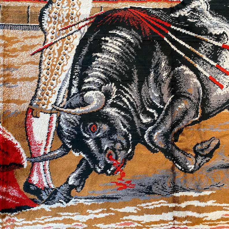 K&ouml;nnte beinhalten: Ein Wandteppich, der eine Stierszenen darstellt. Ein schwarzer Stier mit grauen und wei&szlig;en Akzenten greift eine Figur in einem roten Umhang an. Der Stier hat rote Augen und Blut, das aus seinem Maul tropft. Der Hintergrund hat ein beige-braunes Farbschema.