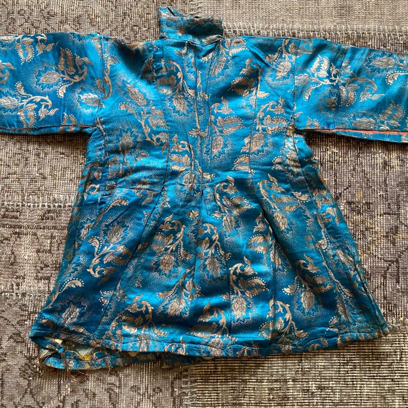 Puede incluir: Una t&uacute;nica de brocado azul y dorado con cuello alto y mangas largas. La tela tiene un patr&oacute;n floral.