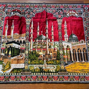 May include: A rectangular prayer rug featuring a detailed depiction of Islamic architecture. The central image shows a mosque with multiple minarets and a green dome, set against a red backdrop. The rug's border is decorated with floral patterns in red, black, and green.