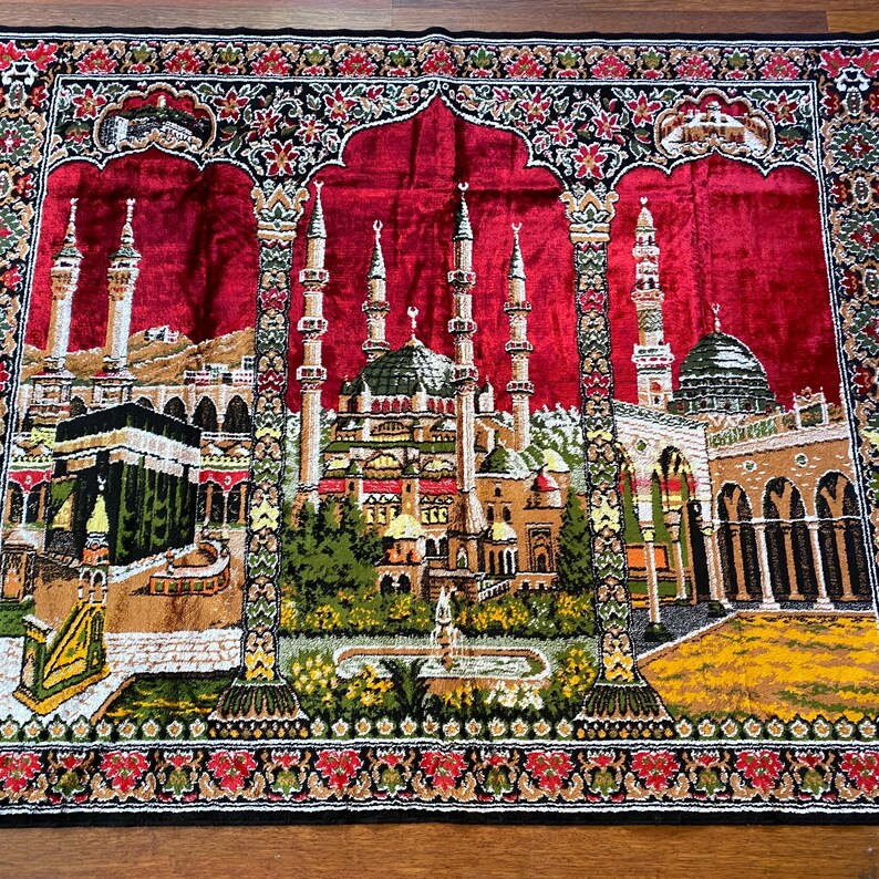 May include: A rectangular prayer rug featuring a detailed depiction of Islamic architecture. The central image shows a mosque with multiple minarets and a green dome, set against a red backdrop. The rug's border is decorated with floral patterns in red, black, and green.