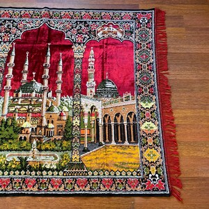 May include: A red prayer mat featuring a detailed mosque design with minarets, domes, and arched doorways. The mat has a decorative border with floral patterns and red fringe.