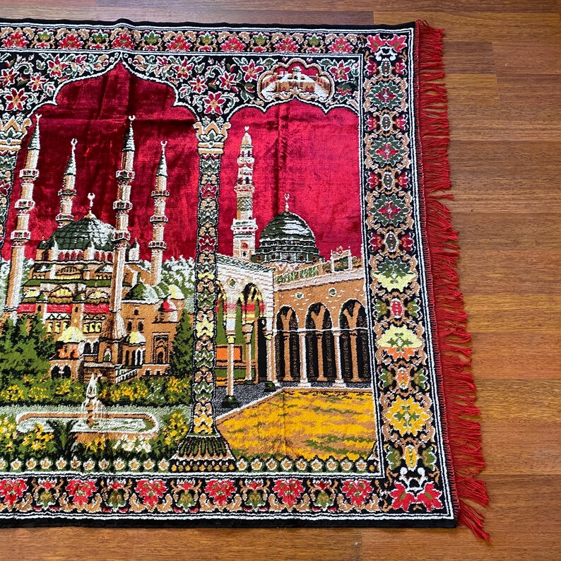 May include: A red prayer mat featuring a detailed mosque design with minarets, domes, and arched doorways. The mat has a decorative border with floral patterns and red fringe.