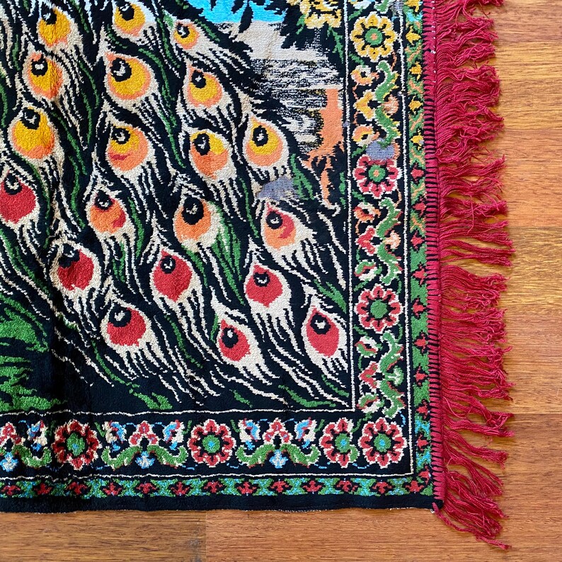 May include: A section of a vintage peacock-themed rug with a black background. The rug features colourful peacock feather designs in red, orange, yellow, and green. The border has floral patterns and red fringe.