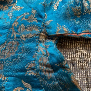 Puede incluir: Primer plano de una tela azul turquesa con un patr&oacute;n floral dorado. La tela tiene un borde deshilachado y est&aacute; ligeramente arrugada.