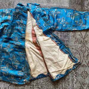 Puede incluir: Una chaqueta de brocado azul y dorado con un patr&oacute;n floral. La chaqueta est&aacute; abierta para revelar un forro blanco.