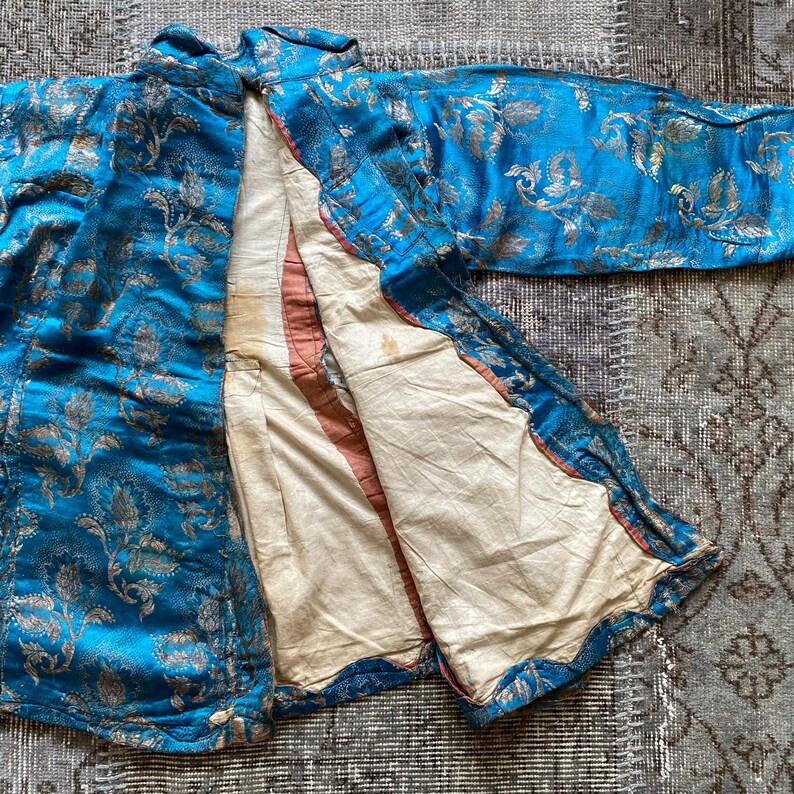 Puede incluir: Una chaqueta de brocado azul y dorado con un patr&oacute;n floral. La chaqueta est&aacute; abierta para revelar un forro blanco.