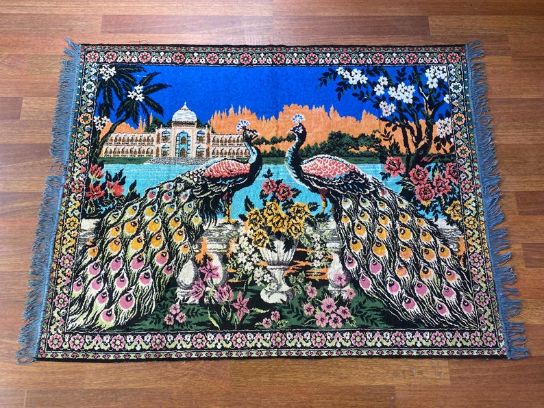 Peut inclure: Une tapisserie rectangulaire repr&eacute;sentant deux paons au plumage vibrant, sur fond de ciel bleu, de lac et de palais. La tapisserie est bord&eacute;e d'un motif floral et a des bords frang&eacute;s. Les paons sont au centre, avec leurs plumes color&eacute;es.
