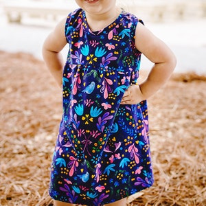 Pode incluir: Um vestido sem mangas para uma menina pequena, com fundo azul escuro e estampa floral e de pássaros colorida.