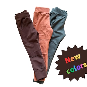 Puede incluir: Tres pares de pantalones de chándal en marrón, coral y gris. Los pantalones de chándal están doblados y apilados uno encima del otro. Un estallido de estrella negro con el texto "Nuevos colores" está en el fondo.