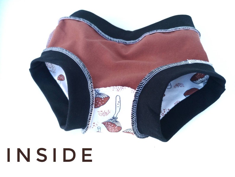 Pode incluir: Roupa interior com um painel de cor ferrugem s&oacute;lida, faixas el&aacute;sticas pretas na cintura e nas pernas com costuras brancas vis&iacute;veis, e um forro interior branco com um padr&atilde;o de ilustra&ccedil;&otilde;es de cogumelos castanhos. A palavra INSIDE est&aacute; impressa em castanho.