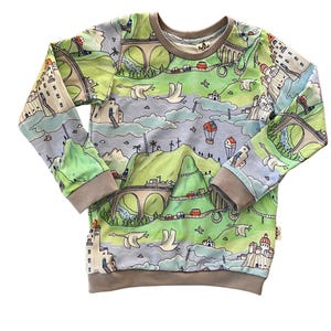 May include: A long-sleeved, light blue sweatshirt with a colourful print of a whimsical landscape featuring mountains, bridges, houses, and birds.
